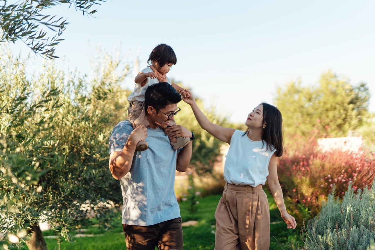 A family enjoys a walk outside. The father carries a child on his shoulders, while the mother reaches out to them. Lush greenery and trees are in the background.